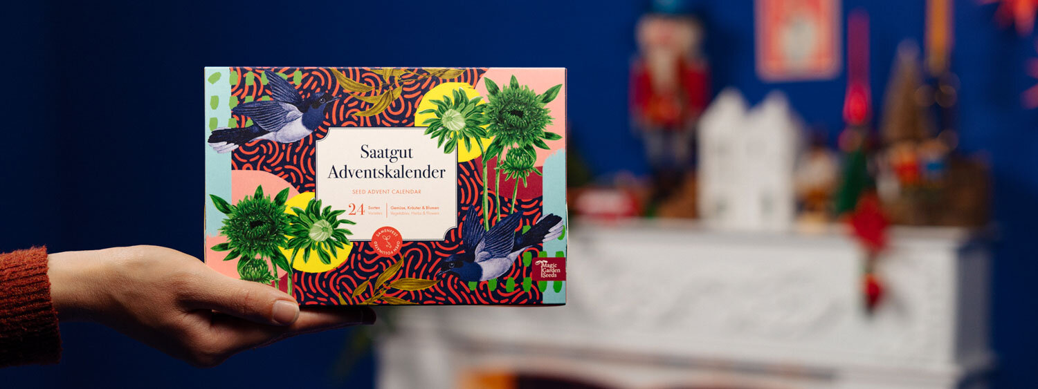 Saatgut-Adventskalender „Gemüse, Kräuter & Blumen“ in der Hand vor weihnachtlich dekoriertem Kamin